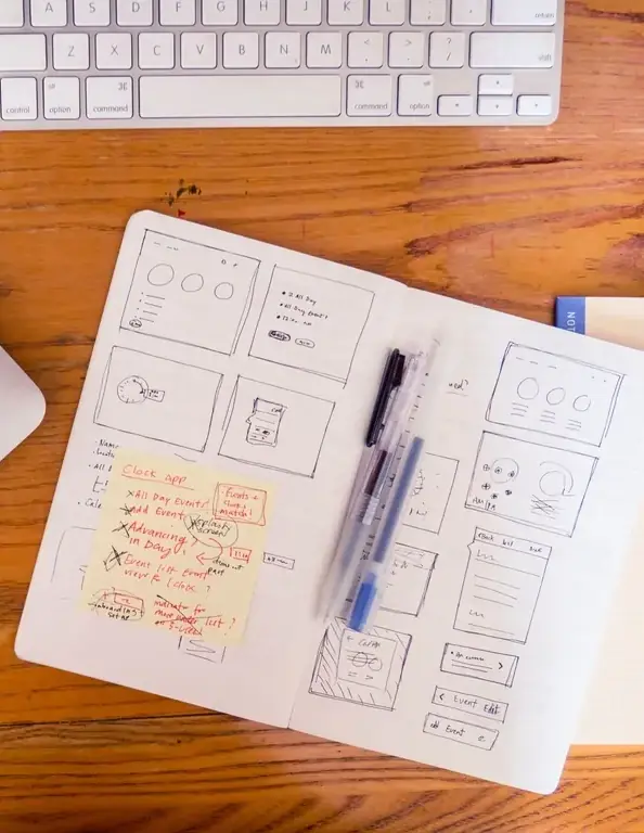 An open sketchbook of hand-drawn screen wireframes with a yellow note of red annotations beside a keyboard